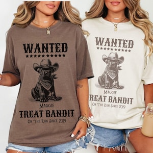 May include: Two t-shirts, one espresso and one ivory, each featuring a graphic of a dog in a cowboy hat and bandana. The text reads "WANTED" above the dog, and "Maggie Treat Bandit On The Run Since 2019" below. The shirts are casual wear.