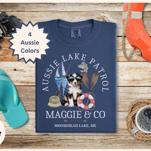 May include: A blue t-shirt with a graphic of an Australian Shepherd dog wearing sunglasses and sitting with fishing gear. The text "Aussie Lake Patrol" is in a circle around the dog. The text "Maggie & Co Moosehead Lake, ME" is below the graphic.