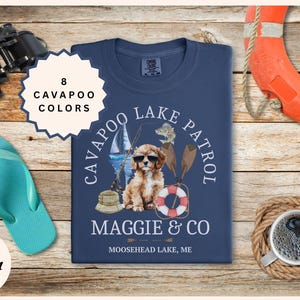 May include: Navy blue t-shirt with a graphic of a Cavapoo dog wearing sunglasses, a sailboat, and the text "Cavapoo Lake Patrol Maggie & Co Moosehead Lake, ME." The shirt is surrounded by beach-themed accessories.