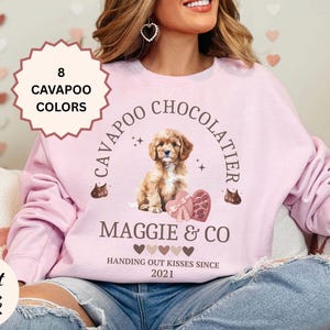 May include: Light pink sweatshirt with a Cavapoo Chocolatier design. The graphic includes a puppy, heart-shaped chocolates, and the text "Maggie & Co. Handing out kisses since 2021." A tag indicates "8 Cavapoo Colors."