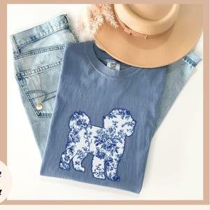 May include: A blue t-shirt with a floral dog design, paired with light blue jeans and a tan hat. The dog graphic is white with blue floral patterns. The words "Blue Jean" are in the bottom left corner.