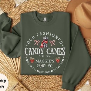 May include: Military green sweatshirt with the text "OLD FASHIONED CANDY CANES" and a dog graphic. The design includes candy canes, paw prints, and the text "MAGGIE'S CANDY CO. EST. 2024".