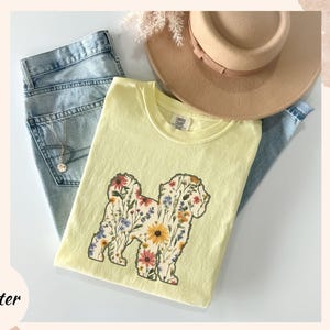 May include: A light yellow t-shirt with a floral dog design, paired with light blue jeans and a tan hat. The dog graphic is filled with colorful flowers. A gold necklace is visible on the jeans.