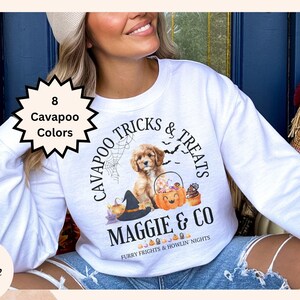May include: A white crewneck sweatshirt featuring a graphic of a Cavapoo puppy, a pumpkin bucket filled with candy, a witch hat, and spiderwebs. The text reads Cavapoo Tricks & Treats, Maggie & Co, and Furry Frights & Howlin' Nights.