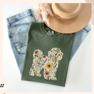 May include: A moss green t-shirt featuring a floral dog design, paired with light blue jeans and a tan hat. The dog silhouette is filled with colorful flowers. The word "Moss" is written in the bottom left corner.