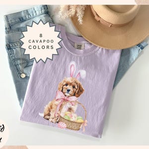 May include: Orchid t-shirt in a light purple color, featuring a Cavalier King Charles Spaniel puppy wearing bunny ears and holding a basket of Easter eggs. The shirt is displayed with jeans and a hat.