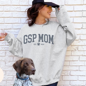 May include: A gray sweatshirt with the text "GSP MOM EST. 2024" and a paw print. A woman is wearing the sweatshirt and holding a glass of wine. A brown and white dog is sitting in front of the woman.
