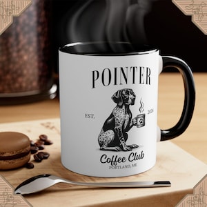 May include: White ceramic coffee mug with a black interior and handle. The mug features a black and white illustration of a Pointer dog holding a coffee cup, with the text "POINTER COFFEE CLUB PORTLAND, ME". A spoon and a macaron are in the foreground.