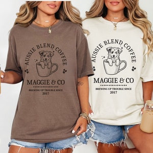 May include: Two women's t-shirts with a brown and white design. The design features a cartoon Australian Shepherd puppy in a coffee cup with the text "Aussie Blend Coffee" and "Maggie & Co. Brewing Up Trouble Since 2017". The shirts are available in espresso and ivory.