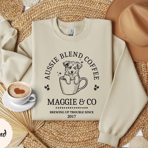 May include: A beige sweatshirt with a black graphic design of a dog in a coffee cup. The text on the graphic reads "Aussie Blend Coffee Maggie & Co Brewing Up Trouble Since 2017".
