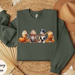 May include: Military green sweatshirt featuring a watercolor design of a Bernese Mountain Dog, pumpkins, fall leaves, and coffee drinks. The sweatshirt is a crew neck style.