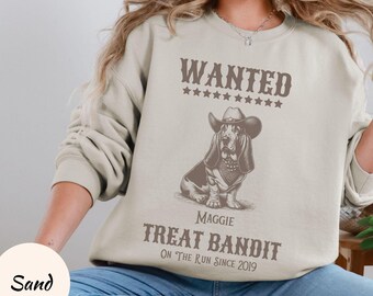 May include: A beige sweatshirt with a graphic of a basset hound wearing a cowboy hat and the text "WANTED" with stars above and "Maggie Treat Bandit On The Run Since 2019" below.