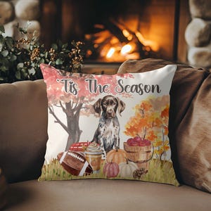 May include: A decorative throw pillow with a fall theme. The pillow features a watercolor illustration of a dog, pumpkins, a football, and the text "Tis The Season". The background includes autumn trees and a wooden bucket filled with apples.