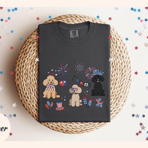 May include: A dark gray t-shirt with a festive Fourth of July design featuring three cartoon poodles wearing red, white, and blue accessories. The poodles are surrounded by fireworks and bunting.