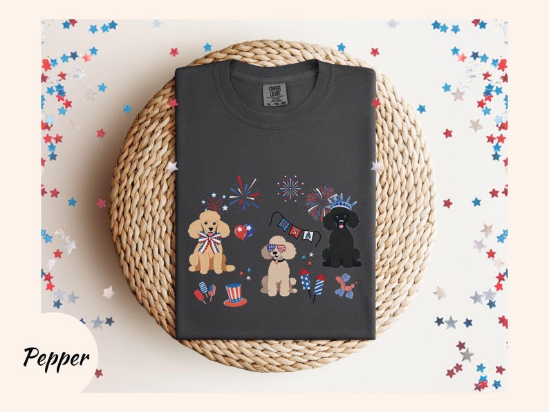 May include: A dark gray t-shirt with a festive Fourth of July design featuring three cartoon poodles wearing red, white, and blue accessories. The poodles are surrounded by fireworks and bunting.
