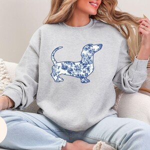 May include: A light gray sweatshirt featuring a blue and white floral dachshund design. The dog illustration is detailed with a floral pattern. The word "Ash" is printed on a light beige circle.