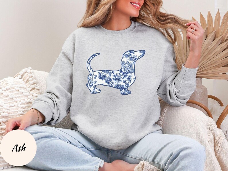 May include: A light gray sweatshirt featuring a blue and white floral dachshund design. The dog illustration is detailed with a floral pattern. The word "Ash" is printed on a light beige circle.