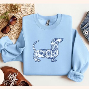 May include: Light blue sweatshirt featuring a dachshund dog silhouette filled with a blue floral pattern. The sweatshirt has a crew neck and long sleeves. The words "Light Blue" are printed in the lower left corner.