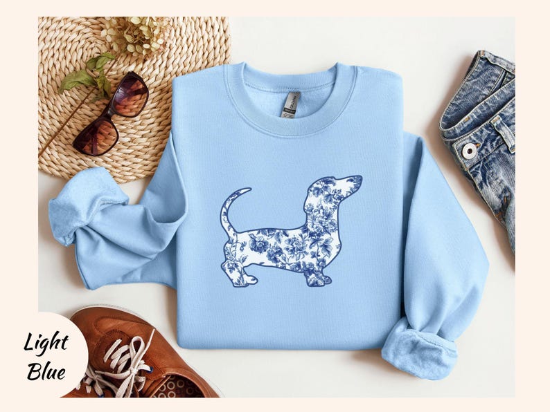 May include: Light blue sweatshirt featuring a dachshund dog silhouette filled with a blue floral pattern. The sweatshirt has a crew neck and long sleeves. The words "Light Blue" are printed in the lower left corner.