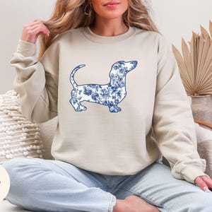 May include: Beige sweatshirt featuring a blue floral dachshund design. The dog illustration is filled with a blue and white floral pattern. The word "Sand" is in a circle in the lower left corner.