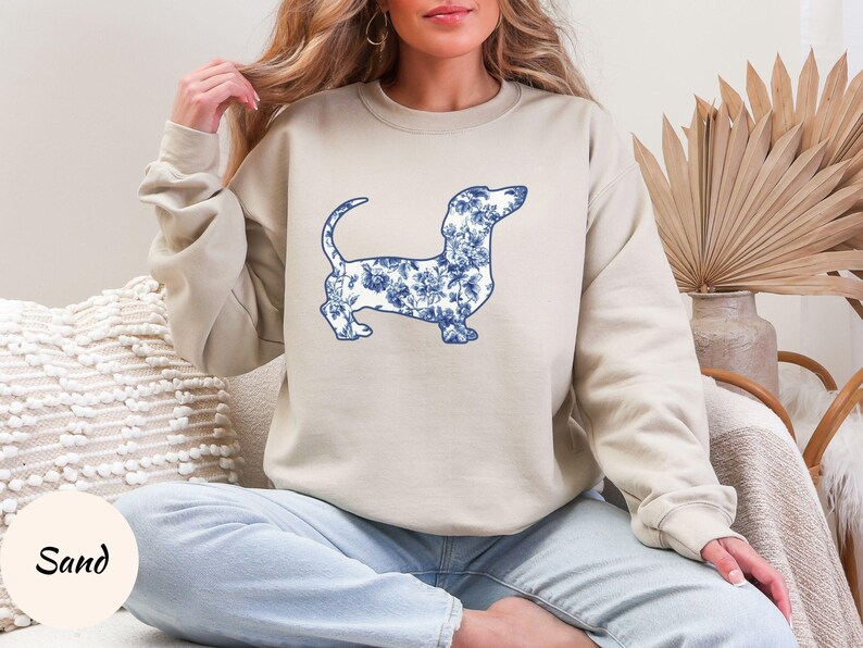 May include: Beige sweatshirt featuring a blue floral dachshund design. The dog illustration is filled with a blue and white floral pattern. The word "Sand" is in a circle in the lower left corner.