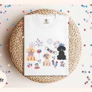 May include: White t-shirt with a festive Fourth of July design featuring three dogs wearing party hats and surrounded by fireworks, stars, and the word "USA".