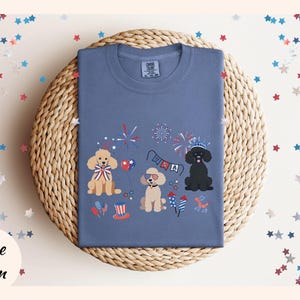 May include: A blue t-shirt with a festive Fourth of July design featuring three dogs wearing red, white, and blue accessories. The dogs are surrounded by fireworks and a banner that says "USA".