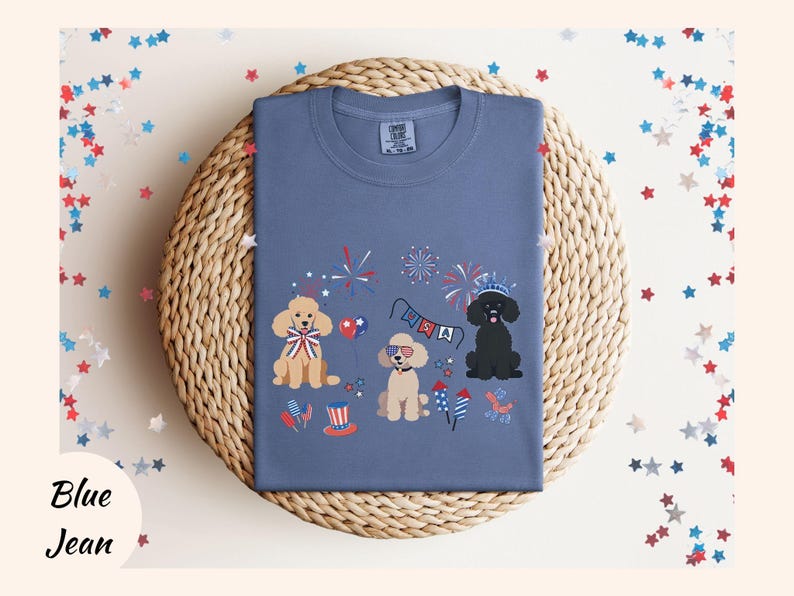 May include: A blue t-shirt with a festive Fourth of July design featuring three dogs wearing red, white, and blue accessories. The dogs are surrounded by fireworks and a banner that says "USA".