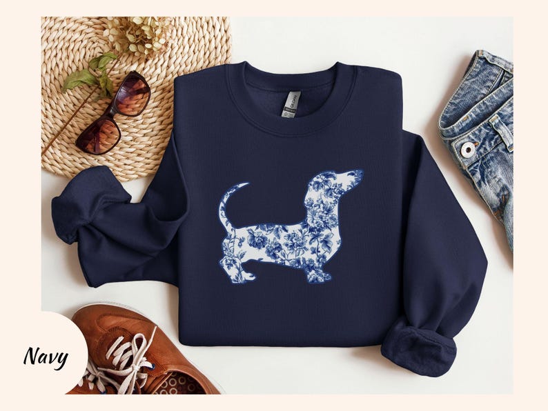 May include: Navy blue sweatshirt featuring a dachshund dog silhouette filled with a blue and white floral pattern. The sweatshirt is displayed with brown shoes, sunglasses, and denim jeans. The word "Navy" is in the bottom left corner.