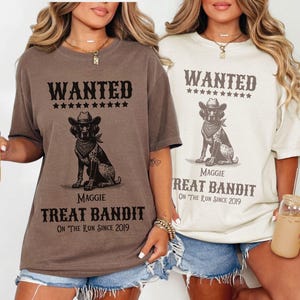 May include: Two brown and cream colored t-shirts with a wanted poster design featuring a dog wearing a cowboy hat. The text on the shirts reads "WANTED" and "Treat Bandit".