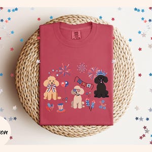 May include: Crimson red t-shirt with a festive Fourth of July design featuring three cartoon poodles wearing patriotic accessories and fireworks. The text "USA" is visible on a banner.