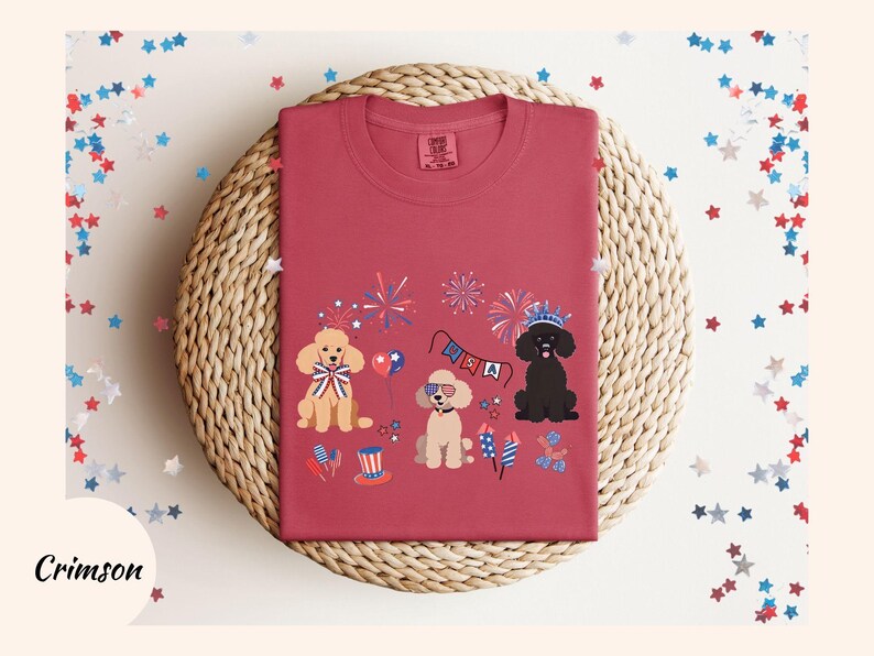 May include: Crimson red t-shirt with a festive Fourth of July design featuring three cartoon poodles wearing patriotic accessories and fireworks. The text "USA" is visible on a banner.