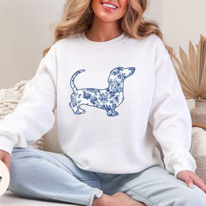 May include: White sweatshirt featuring a blue floral dachshund design. The dog silhouette is filled with a blue and white floral pattern. The sweatshirt has long sleeves and a crew neck. The word "White" is in a circle in the lower left corner.