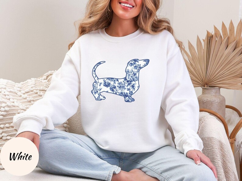May include: White sweatshirt featuring a blue floral dachshund design. The dog silhouette is filled with a blue and white floral pattern. The sweatshirt has long sleeves and a crew neck. The word "White" is in a circle in the lower left corner.