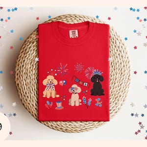May include: Red t-shirt with a graphic of three dogs wearing party hats and celebrating with fireworks and a banner that says "USA".