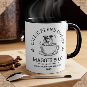May include: A black and white ceramic coffee mug with a black handle. The mug has a design of a Collie dog in a coffee cup with the text "Collie Blend Coffee Maggie & Co Brewing Up Trouble Since 2017".