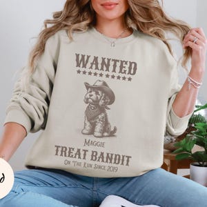 May include: A light beige sweatshirt with a graphic of a dog wearing a cowboy hat and bandana. The text "WANTED" and "TREAT BANDIT" are above and below the dog illustration. The text "ON THE RUN SINCE 2019" is below the dog.