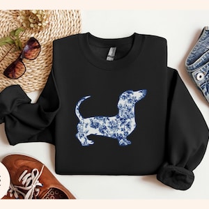 May include: Black sweatshirt featuring a dachshund dog silhouette filled with a blue and white floral pattern. The sweatshirt has long sleeves and a crew neck. The word "Black" is printed on the bottom left.