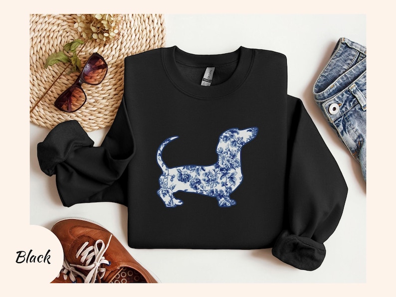 May include: Black sweatshirt featuring a dachshund dog silhouette filled with a blue and white floral pattern. The sweatshirt has long sleeves and a crew neck. The word "Black" is printed on the bottom left.