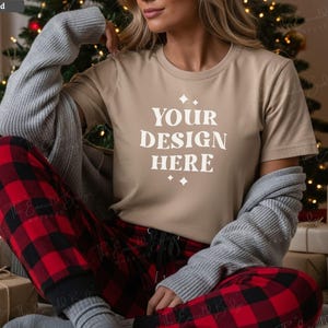 May include: A tan t-shirt with the text "YOUR DESIGN HERE" in white, paired with red and black plaid pajama pants. A gray sweater is draped over the shoulders. The scene is set in front of a Christmas tree.