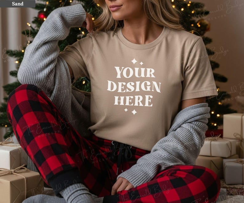 May include: A tan t-shirt with the text "YOUR DESIGN HERE" in white, paired with red and black plaid pajama pants. A gray sweater is draped over the shoulders. The scene is set in front of a Christmas tree.