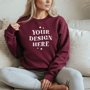 May include: A maroon sweatshirt with the text "YOUR DESIGN HERE" in white lettering. The sweatshirt is being worn by a person sitting on a couch.