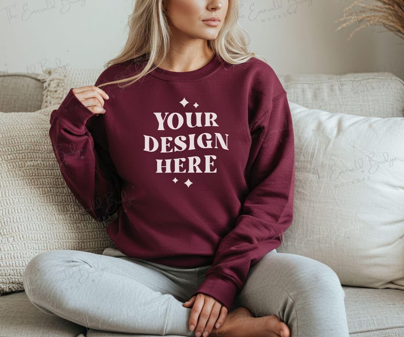 May include: A maroon sweatshirt with the text "YOUR DESIGN HERE" in white lettering. The sweatshirt is being worn by a person sitting on a couch.