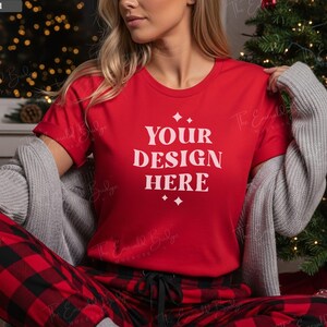 May include: A red t-shirt with the text "YOUR DESIGN HERE" in white, paired with red and black plaid pajama pants. A gray sweater is draped over the shoulders. The scene is set near a Christmas tree.