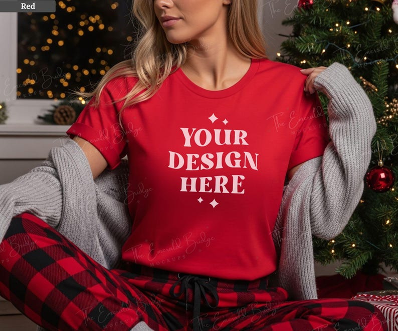 May include: A red t-shirt with the text "YOUR DESIGN HERE" in white, paired with red and black plaid pajama pants. A gray sweater is draped over the shoulders. The scene is set near a Christmas tree.