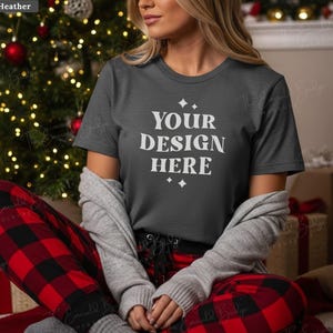 May include: A dark heather gray t-shirt with the text "YOUR DESIGN HERE" in white. The person is wearing red and black plaid pajama pants and a gray cardigan. A Christmas tree with red ornaments and lights is in the background.