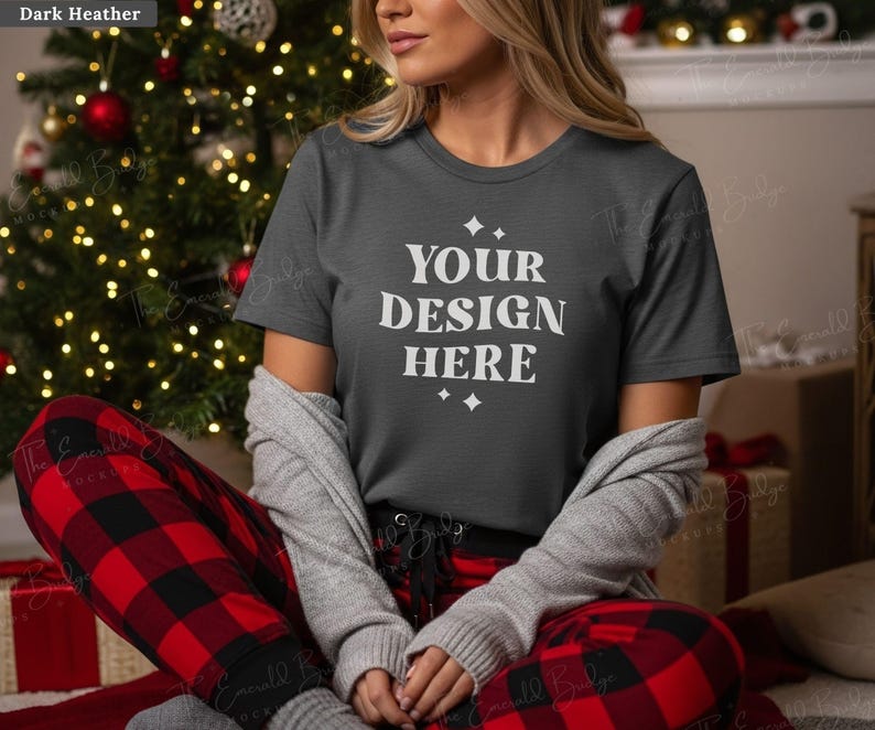 May include: A dark heather gray t-shirt with the text "YOUR DESIGN HERE" in white. The person is wearing red and black plaid pajama pants and a gray cardigan. A Christmas tree with red ornaments and lights is in the background.