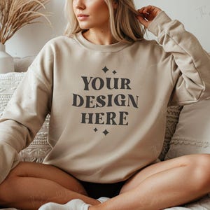 May include: A beige sweatshirt with the text "YOUR DESIGN HERE" printed in black on the front. The sweatshirt is being worn by a person sitting on a couch.