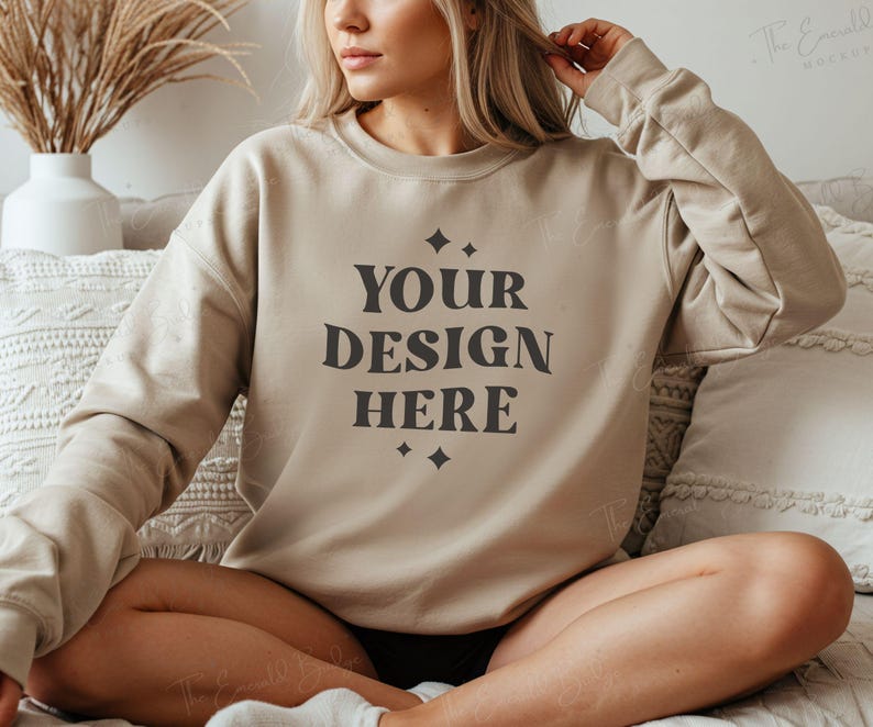 May include: A beige sweatshirt with the text "YOUR DESIGN HERE" printed in black on the front. The sweatshirt is being worn by a person sitting on a couch.