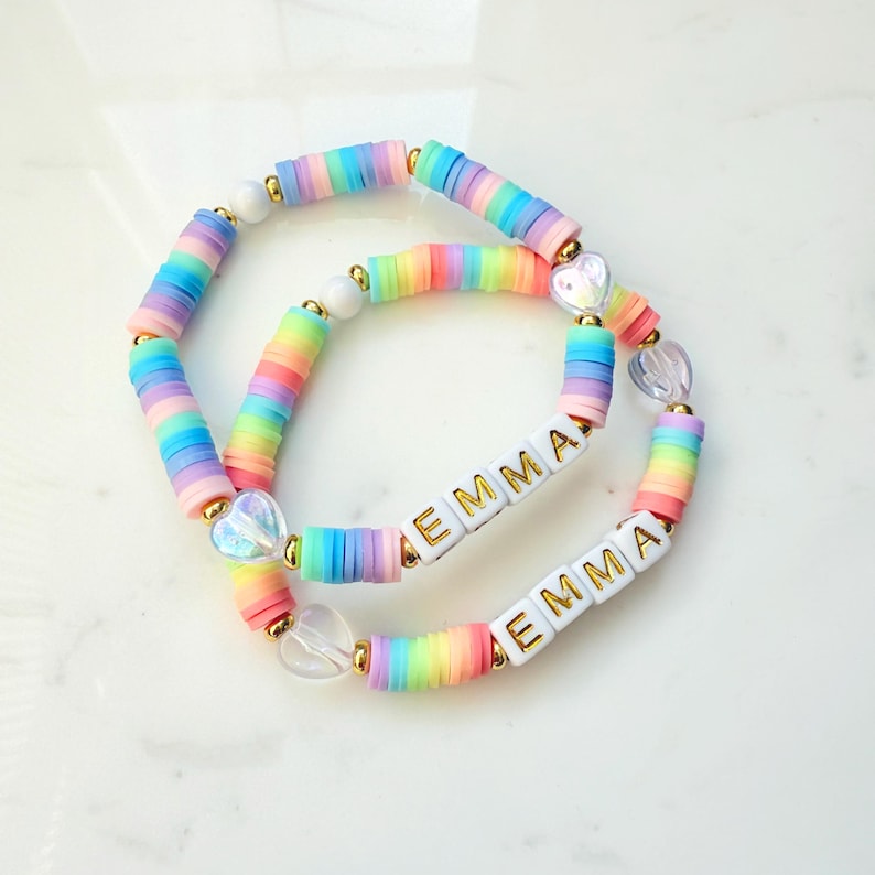 May include: Two bracelets with rainbow-coloured disc beads and gold accents. One bracelet features the name "EMMA" spelled out in gold letters on white beads. Clear heart-shaped charms and white beads are also included.